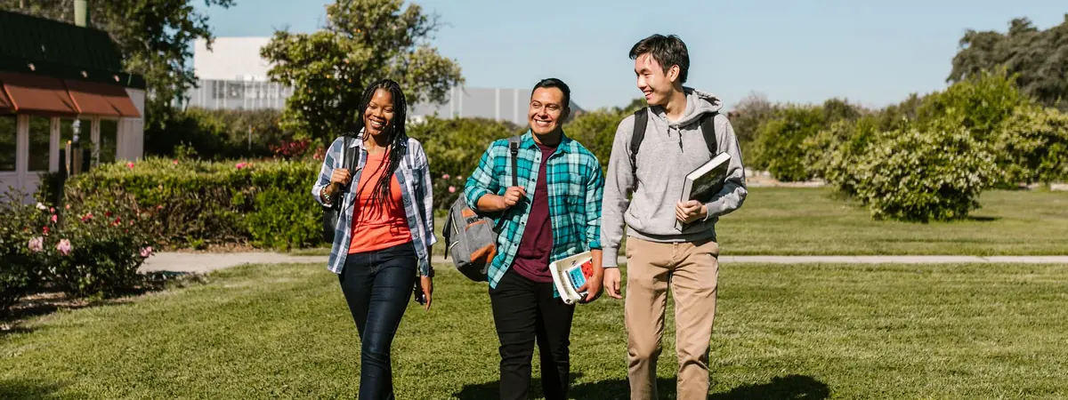 Three college students carry backpacks and textbooks across a campus lawn, starting an associate degree.