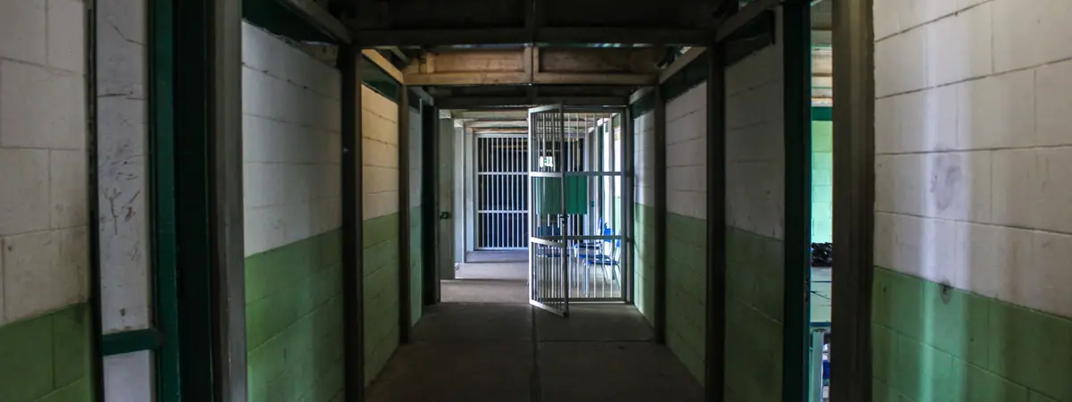 An empty prison corridor with green-and-white block walls leads to an open barred door, a correctional officer’s workplace.