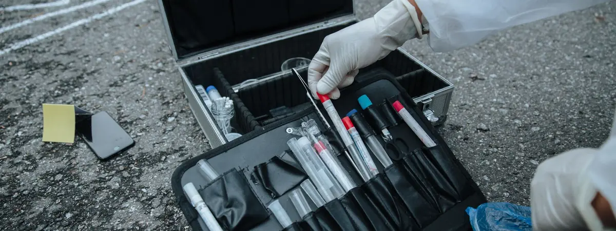 A gloved hand lifts a tube from an evidence kit beside chalk marks, crime scene investigator work.