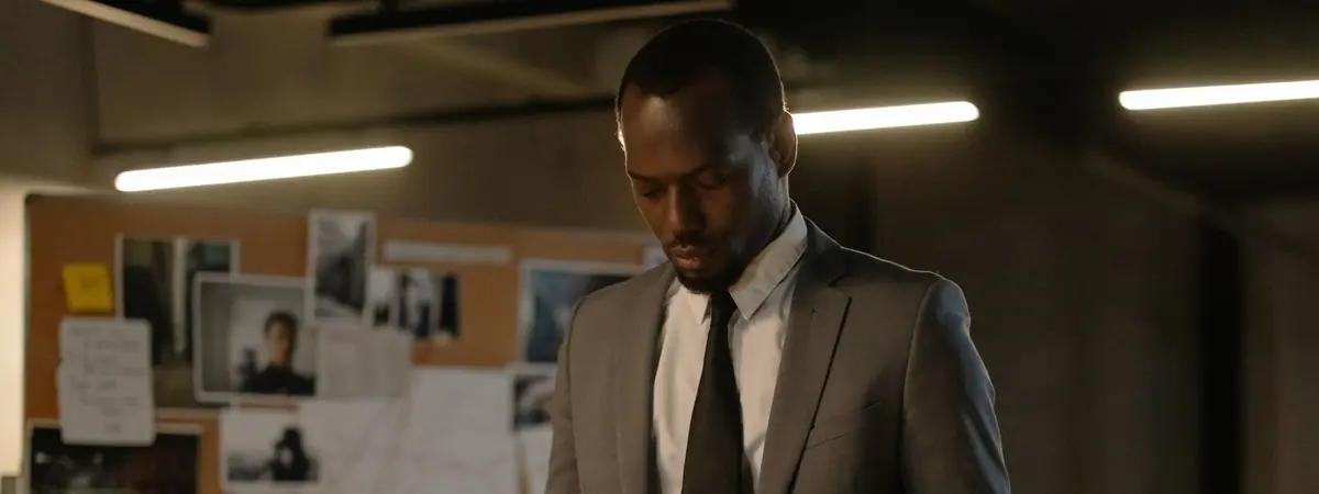 A man in a gray suit stands before a corkboard of pinned photos, the work of a detective.