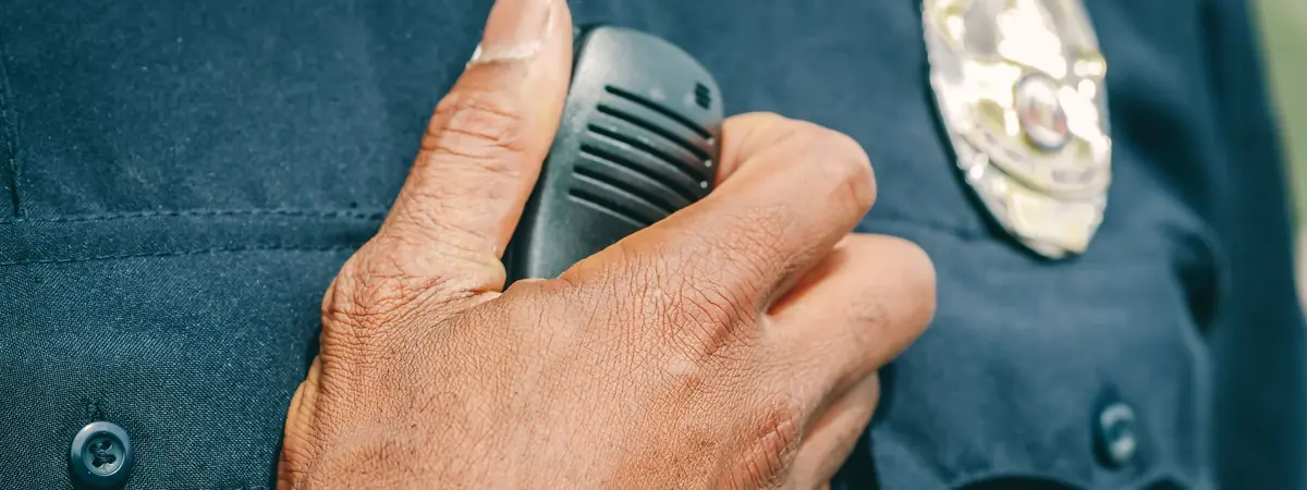 A hand grips a radio on a dark police uniform shirt, the gear behind becoming a police officer.