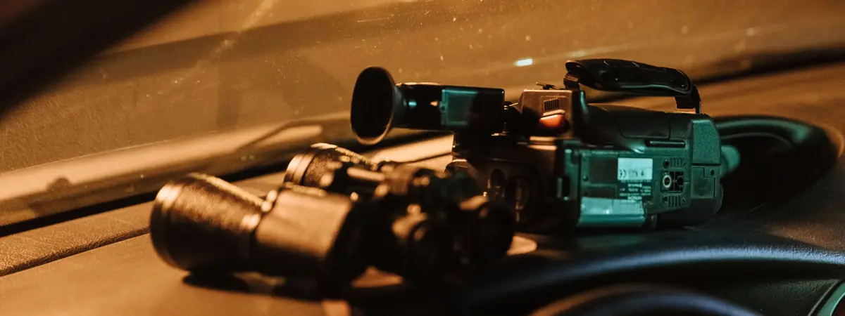 A camcorder and binoculars rest on the dashboard of a parked car, tools a private investigator might use.