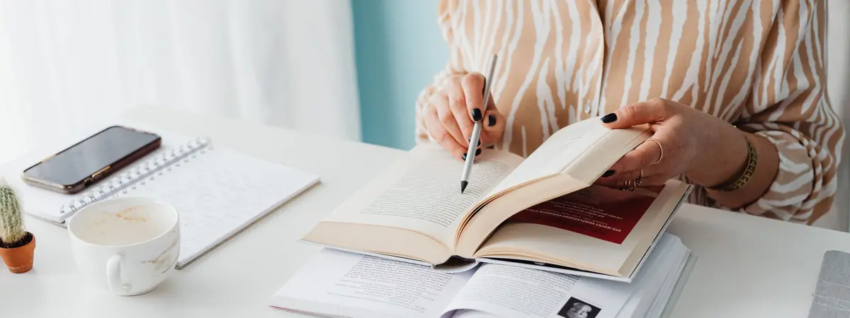 A woman reads an open book with a pen in hand, weighing criminology vs criminal justice.