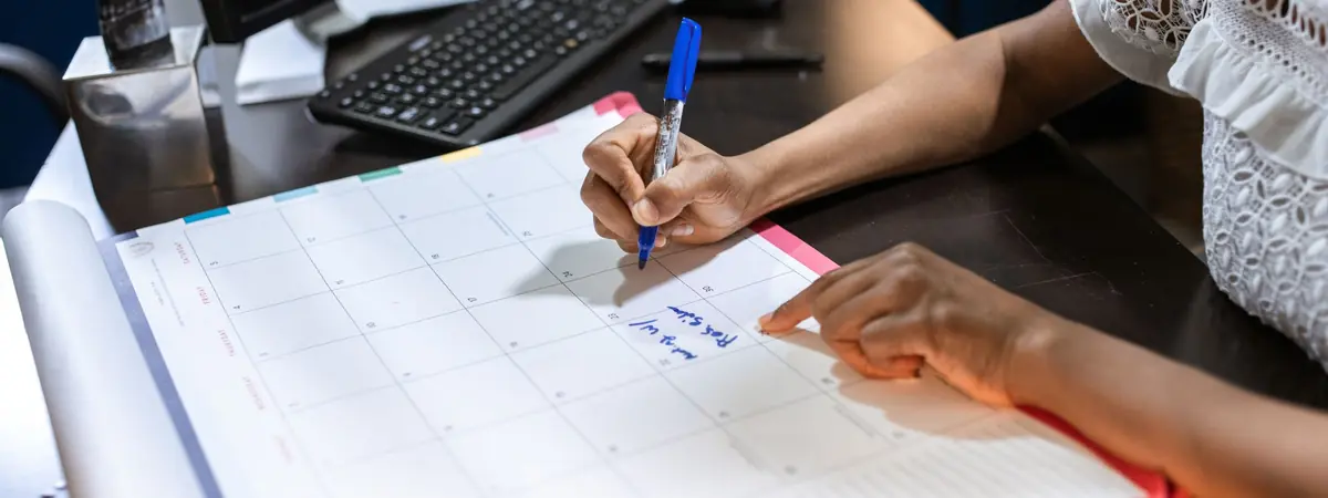 A hand writes in a monthly desk planner, scheduling an accelerated online criminal justice degree.